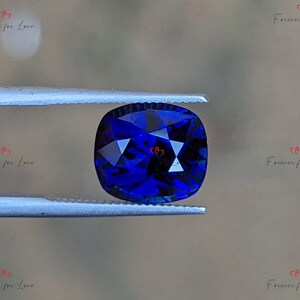 May include: A deep blue cushion-cut gemstone held in tweezers. The gemstone has a faceted surface and is a vibrant blue color.