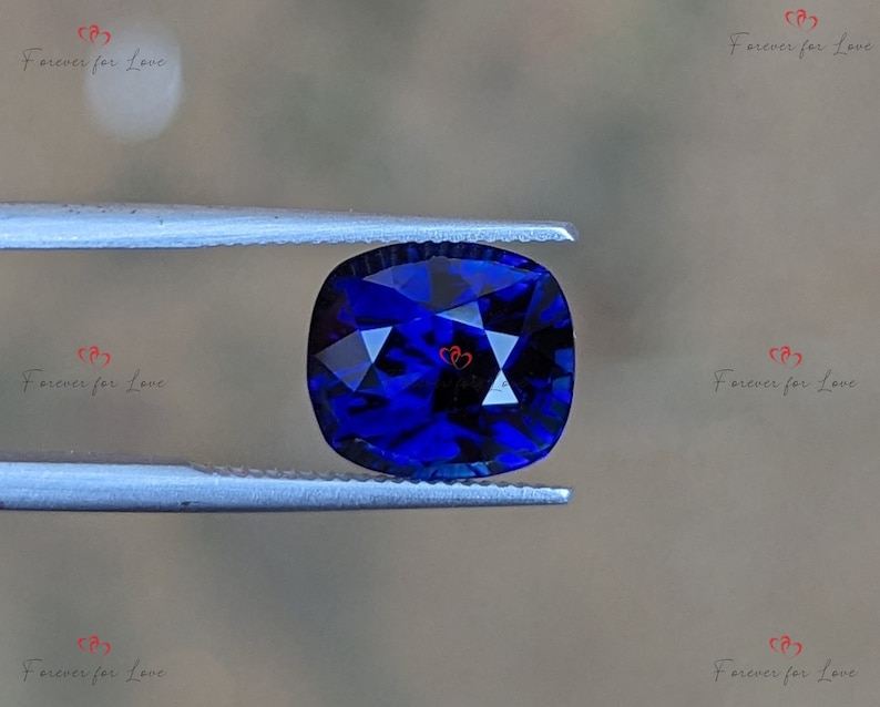 May include: A deep blue cushion-cut gemstone held in tweezers. The gemstone has a faceted surface and is a vibrant blue color.