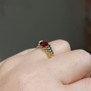Puede incluir: Un anillo de oro con una gran gema roja ovalada. El anillo tiene una banda texturizada con peque&ntilde;as piedras de acento. El dise&ntilde;o es elegante y cl&aacute;sico, adecuado para ocasiones especiales.