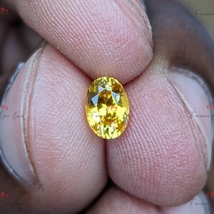 May include: A yellow oval-shaped gemstone with facets. The gemstone is held between two fingers.