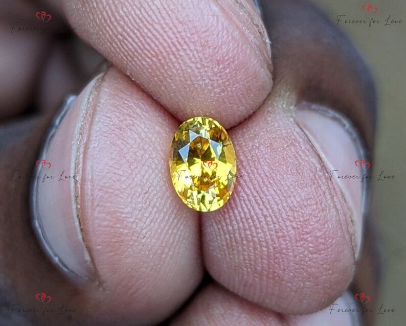 May include: A yellow oval-shaped gemstone with facets. The gemstone is held between two fingers.