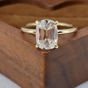 Może przedstawiać: Złoty pierścionek zaręczynowy z dużą, prostokątną, przezroczystą szlachetnym kamieniem. Pierścionek leży na drewnianym pudełku z ciemnobrązowym wykończeniem. Kamień szlachetny jest utrzymywany przez cztery pazury.