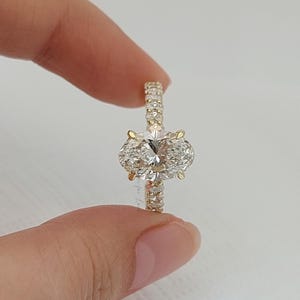 May include: A gold engagement ring featuring a large, oval-cut diamond as the centerpiece. The band is adorned with smaller, sparkling diamonds. The ring is held up against a plain background, showcasing its brilliance and design.