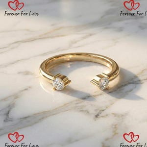 Könnte beinhalten: Ein goldfarbener Ring mit zwei klaren, runden Edelsteinen an den offenen Enden. Der Ring hat ein glattes, poliertes Band und wird auf einer Marmoroberfläche präsentiert. Der Text "Forever For Love" ist im Hintergrund sichtbar.