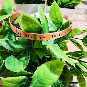 May include: A gold-toned metal bracelet with the words "You are my sunshine" engraved on it. The bracelet is surrounded by green leaves.