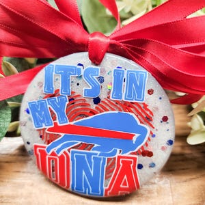 May include: A circular ornament with the text "IT'S IN MY DNA" in blue and red. The ornament features a blue and red buffalo logo and is tied with a red ribbon.