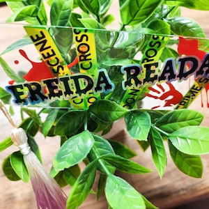 May include: A clear acrylic tag with the name "FREIDA READAH" in black and blue lettering, accented with yellow "SCENE CROSS" and "CRIME SCENE" tape. Red blood splatter graphics are also present. The tag is attached to a green leafy plant.
