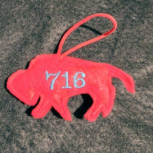 Op de afbeelding: Rode vilten buffel ornament met een blauw geborduurd nummer "716" aan de zijkant.
