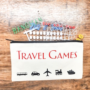 May include: A white canvas travel games pouch with a black zip, red text, and black illustrations of transport. The pouch contains game pieces and the words "HANGMAN" and "Days" in green and blue.