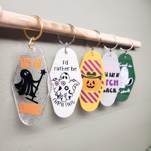 May include: A collection of Halloween-themed keychains hanging on a wooden dowel. The keychains feature various designs, including ghosts, pumpkins, and phrases like "Let's Go Ghouls" and "I'd rather be thrifting." Each keychain has a metal ring.