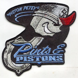 Op de afbeelding: Een zwarte geborduurde patch met een cartoonfiguur met een hoed en een hoefijzer. De tekst "Pints & Pistons" is in blauw geborduurd op de patch.