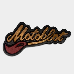 May include: A black and gold leather patch with the word "Motoblott" in a stylised font. The patch has a red flame design at the bottom.