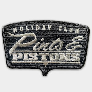 May include: Black embroidered patch with white stitching. The patch features the text "Holiday Club" at the top, "Pints & Pistons" in the centre, and a pointed bottom.