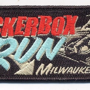 May include: A brown embroidered patch with the text "Rockerbox Run 2008 Chicago Milwaukee" in white, red, and blue lettering. A stylised image of a motorcycle rider is in the background.