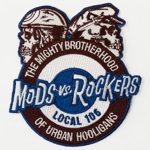 May include: A brown and white embroidered patch featuring two skull heads wearing helmets. The patch reads "The Mighty Brotherhood Mods vs Rockers Local 106 Of Urban Hooligans".