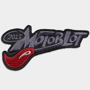 Może przedstawiać: Naszywka haftowana w kolorze czarnym i szarym z tekstem "MotorBlot" w stylizowanym kroju pisma. Naszywka zawiera również wzór płomienia w kolorze czerwonym i białym oraz rok "2015" w kółku.