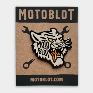 May include: A pin featuring a stylized tiger head with the text "MOTOBLOT" in a bold font. The pin is black, white, and orange, mounted on a brown card with the words "MOTOBLOT" and "MOTOBLOT.COM".