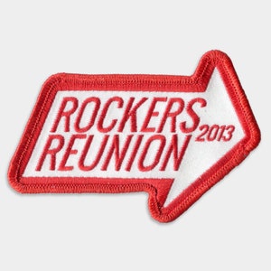 Op de afbeelding: Rode en witte geborduurde patch met de tekst "Rockers Reunion 2013" in de vorm van een witte pijl.