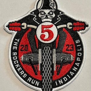 Może przedstawiać: Naszywka haftowana w kolorze czarnym i białym z czerwonym tłem. Naszywka przedstawia szkielet jadący na motocyklu z numerem 5 z przodu. Tekst "The Rockers Run 2023 Indianapolis" znajduje się wokół krawędzi naszywki.