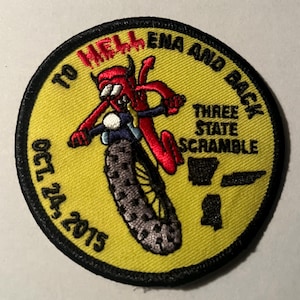 May include: A yellow round patch with black stitching. The patch features a cartoon red devil riding a motorcycle with the text "To Hellena and Back" and "Three State Scramble" around the edge. The date "Oct 24, 2015" is printed on the bottom left side of the patch.