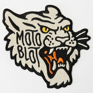 May include: White embroidered tiger head patch with black outline and orange details. The text "Moto Blot" is embroidered on the tiger's face.