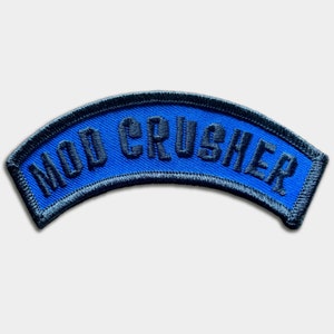 May include: A blue and black embroidered patch with the text "MOD CRUSHER" in black.