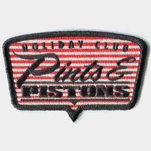 May include: A black and white embroidered patch with red and white stripes. The patch says "Holiday Club Pinto's Pistons".