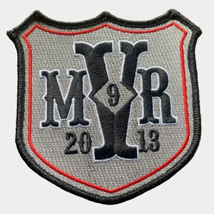 Könnte beinhalten: Ein grauer gestickter Aufnäher in Form eines Schildes mit schwarzem Rand und roten Nähten. Der Aufnäher zeigt die Buchstaben "MYR" in einer stilisierten Schriftart mit einer Diamantenform, die die Zahl "9" in der Mitte enthält. Die Jahre "2013" sind ebenfalls auf den Aufnäher gestickt.