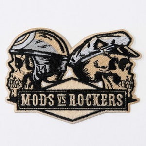 May include: Embroidered patch featuring two skull heads wearing helmets, one with a motorcycle helmet and the other with a military style cap. The patch has a beige background with black stitching and the text "MODS VS ROCKERS" in a black banner.
