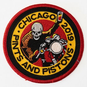 May include: A round, red, black, and yellow embroidered patch with a skeleton riding a motorcycle and the text "Chicago Pints and Pistons 2019."