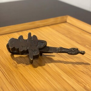 May include: A bronze-colored metal key with an ornate design, resting on a bamboo tray. The key has a detailed handle and a decorative top. The background is a dark surface.