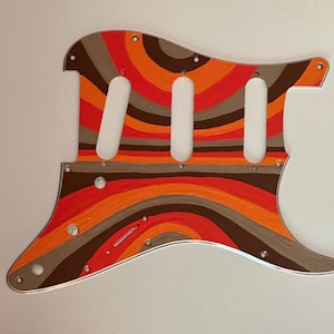 May include: A guitar pickguard featuring a multi-colored pattern of concentric, wavy stripes in shades of bright orange, dark brown, and light gray. The flat, contoured plastic component includes three elongated pickup cutouts and eleven small screw holes.
