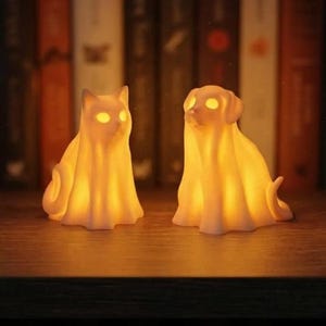 May include: Two small, glowing, off-white figurines shaped like a cat and a dog, both draped in ghost-like sheets. The figures emit a warm, internal amber light from their eyes and bodies, creating a soft illumination against a dark background.