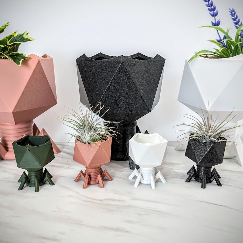 May include: Six geometric planter pots in various colors, including black, white, pink, and green. The pots are shaped like a geometric figure with legs that resemble a spider. The pots are filled with small plants.