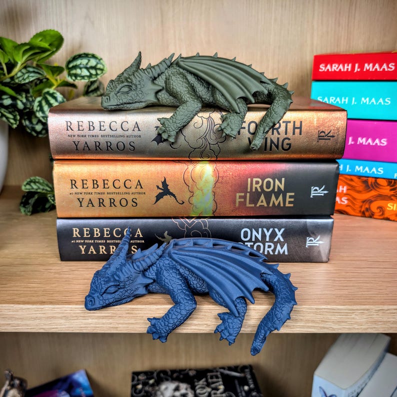 Puede incluir: Una estanter&iacute;a de madera con libros de fantas&iacute;a de Rebecca Yarros y Sarah J. Maas. Una figura de drag&oacute;n verde descansa sobre los libros, con un drag&oacute;n azul debajo. Los t&iacute;tulos incluyen "Fourth Wing", "Iron Flame" y "Onyx Storm".