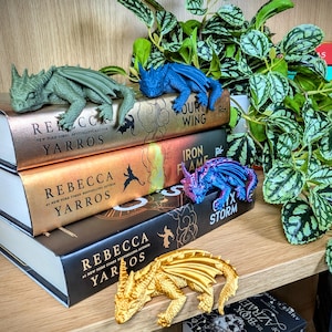 Puede incluir: Una colecci&oacute;n de libros de fantas&iacute;a con figuras de dragones. Los libros tienen cubiertas doradas y negras con el nombre de la autora, Rebecca Yarros, y t&iacute;tulos visibles. Cuatro figuras de dragones en verde, azul, morado y dorado est&aacute;n sobre los libros.