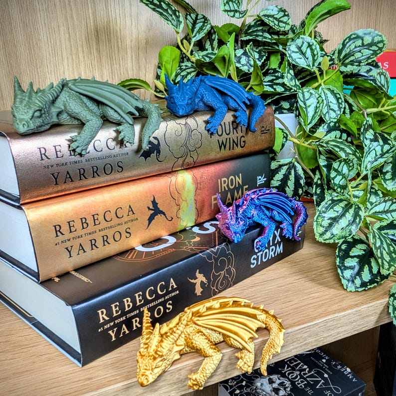 Puede incluir: Una colecci&oacute;n de libros de fantas&iacute;a con figuras de dragones. Los libros tienen cubiertas doradas y negras con el nombre de la autora, Rebecca Yarros, y t&iacute;tulos visibles. Cuatro figuras de dragones en verde, azul, morado y dorado est&aacute;n sobre los libros.