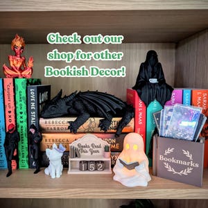 May include: A collection of bookish decor items including a red flame figure, a black dragon, a black hooded figure, a white cat with a purple butterfly, a ghost reading a book, a book tracker with the text Books Read This Year, and a bookmark holder.
