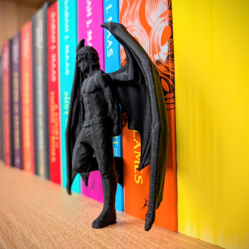 Puede incluir: Una figura negra impresa en 3D de un personaje humanoide alado de pie en perfil. La figura presenta musculatura detallada, piel texturizada y grandes alas extendidas hacia afuera, colocada entre los lomos de dos libros coloridos.