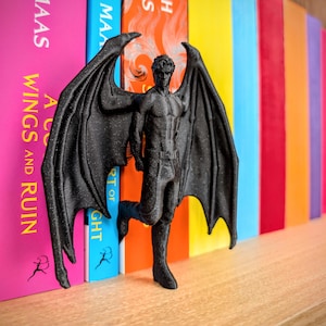 Puede incluir: Una figura negra texturizada de un ser alado con complexi&oacute;n musculosa, de pie con grandes y detalladas alas similares a las de un murci&eacute;lago extendidas. La figura lleva pantalones con un cintur&oacute;n visible y est&aacute; situada entre dos lomos de libros.