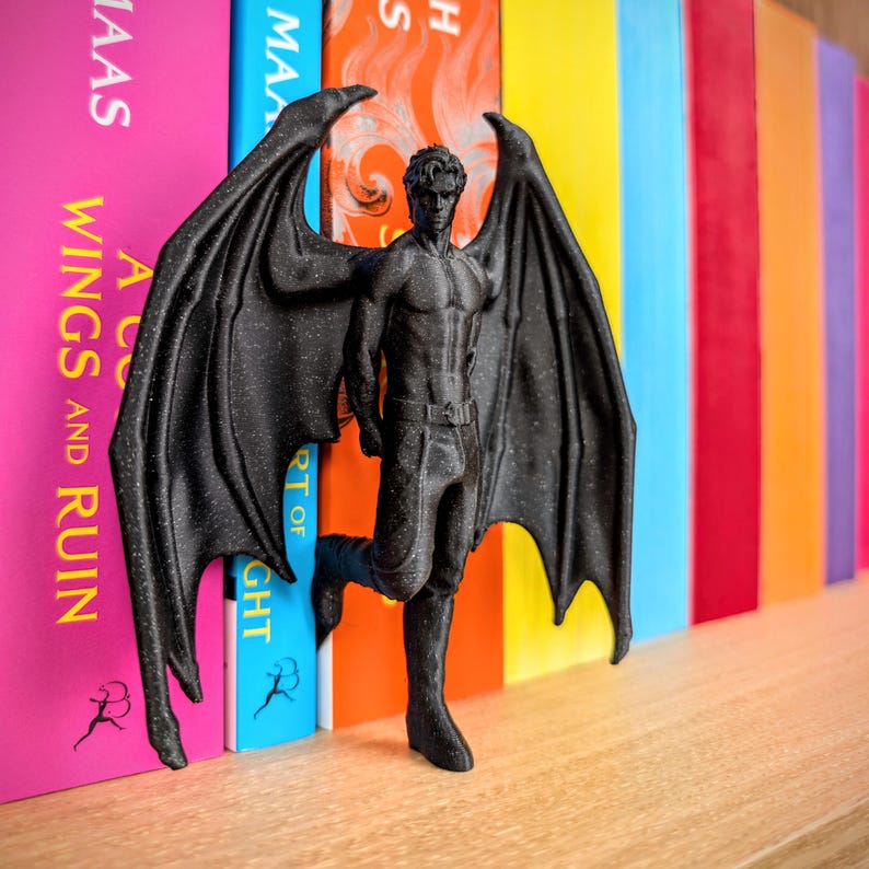 Puede incluir: Una figura negra texturizada de un ser alado con complexi&oacute;n musculosa, de pie con grandes y detalladas alas similares a las de un murci&eacute;lago extendidas. La figura lleva pantalones con un cintur&oacute;n visible y est&aacute; situada entre dos lomos de libros.