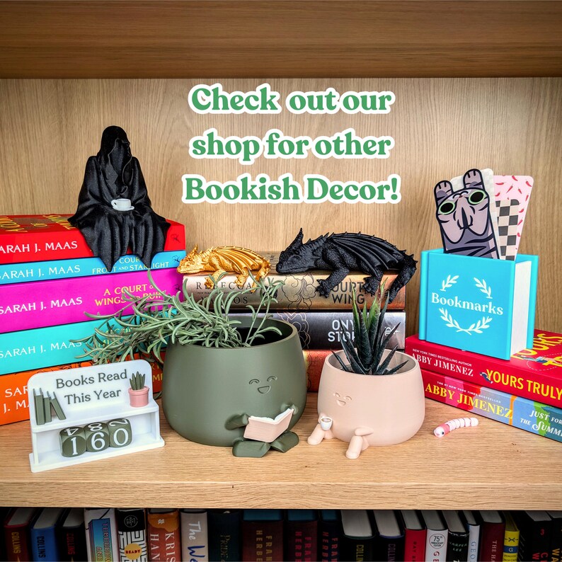 Puede incluir: Una estanter&iacute;a de madera exhibe varios elementos decorativos con tem&aacute;tica de libros. Hay una figura negra de la Parca, figuras de dragones y macetas con caras. Una peque&ntilde;a estanter&iacute;a blanca muestra "Books Read This Year" con un contador en 160. El texto "Check out our shop for other Bookish Decor!"