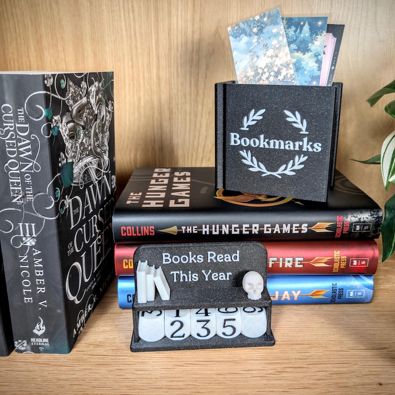 Peut inclure: Une bo&icirc;te noire avec l'inscription "Bookmarks" et un motif de couronne de laurier, contenant des marque-pages d&eacute;coratifs. Un petit pr&eacute;sentoir indique "Livres lus cette ann&eacute;e" avec un cr&acirc;ne et un affichage num&eacute;rique "235". Plusieurs livres en arri&egrave;re-plan.