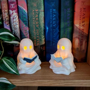 May include: Two white ghost-shaped lamps with glowing yellow eyes, each holding a small book. The left ghost holds a dark blue book, while the right ghost holds a white book. The figures are positioned on a wooden surface in front of seven books.