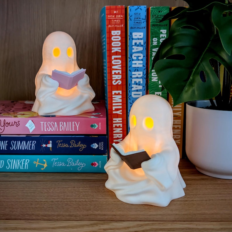 May include: Two white ghost-shaped lamps with glowing yellow eyes, each holding a small book. One ghost holds a pink book, and the other holds a dark brown book. They are positioned on a wooden surface in front of a stack of colorful books.