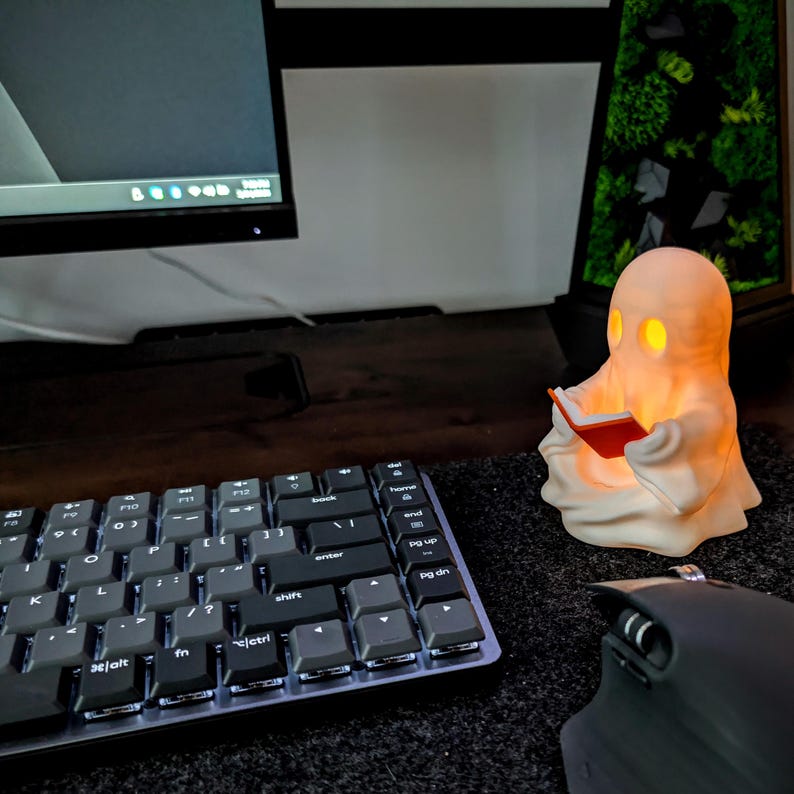 May include: A small, off-white, ghost-shaped lamp with glowing yellow eyes and mouth, holding a miniature red book. The ghost figure is positioned on a dark surface next to a black computer keyboard and a black computer mouse.