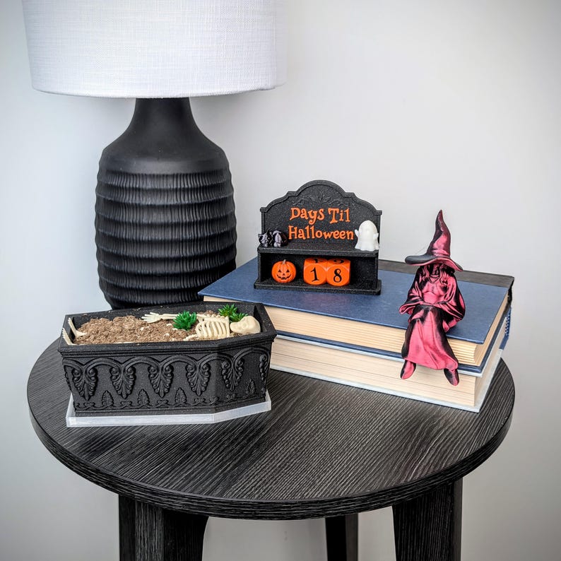 May include: A black coffin-shaped planter containing a skeleton and green succulents, a black Days Til Halloween countdown sign with orange blocks showing 18, and a red witch figurine sitting on a stack of two books with blue covers.