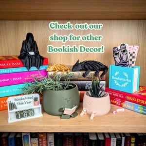 Peut inclure: Une &eacute;tag&egrave;re en bois pr&eacute;sente des d&eacute;corations sur le th&egrave;me des livres. On y trouve une figurine noire de la Faucheuse, des figurines de dragons et des pots de fleurs avec des visages. Une petite &eacute;tag&egrave;re blanche indique "Livres lus cette ann&eacute;e" avec le chiffre 160.