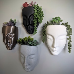 May include: Four different face-shaped planters, each with a different colour and design. The planters are made of ceramic and have a smooth finish. Each planter has a small plant growing inside.