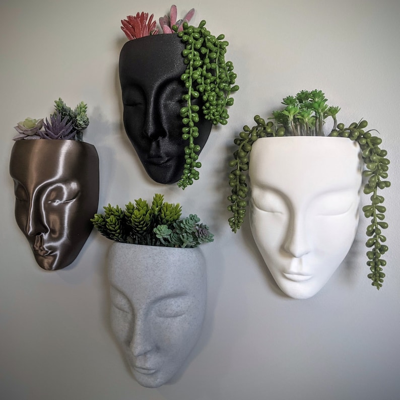 May include: Four different face-shaped planters, each with a different colour and design. The planters are made of ceramic and have a smooth finish. Each planter has a small plant growing inside.